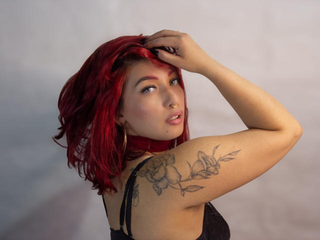BriielaEva con i capelli rossi, che guarda indietro, con una mano tra i capelli, top di pizzo nero, tatuaggio di una rosa sul braccio destro, orecchini a cerchio, un leggero sorriso, seno di media grandezza, pelle chiara, luce soffusa..
