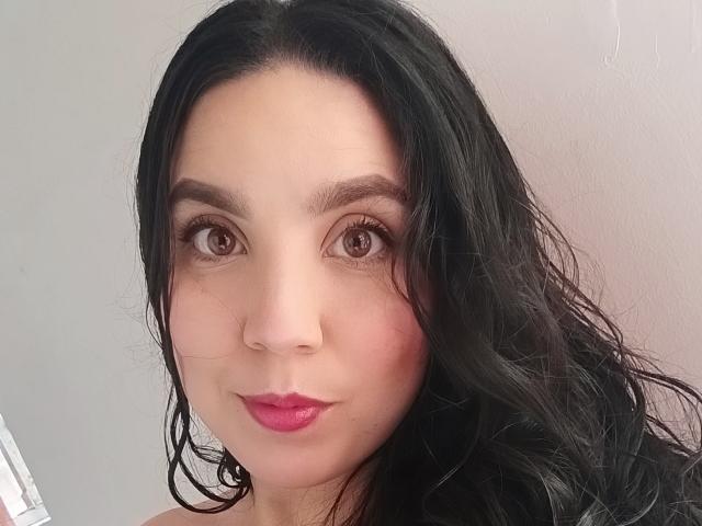 Amelia Gacio tiene el cabello oscuro y ondulado, ojos marrones y lápiz labial rosa. Su rostro ligeramente sonrojado y su sonrisa sutil están en primer plano. Está mirando directamente a la cámara..