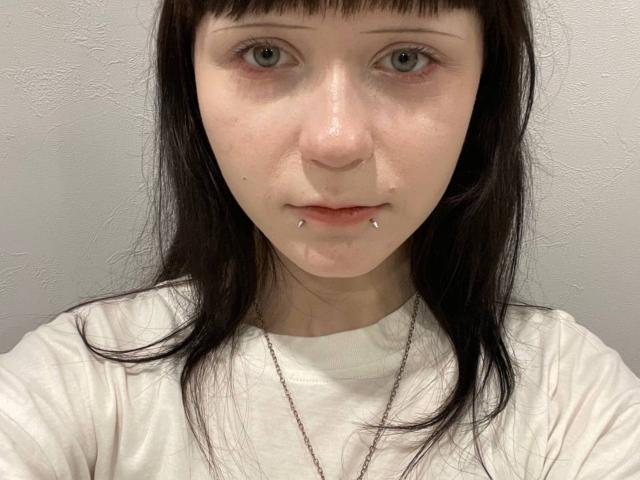 Nancy Turpin, met bleke huid, groene ogen, donkerbruin steil haar, een wit T-shirt en een piercing in de onderlip, draagt een dunne ketting, op een eenvoudige selfie..