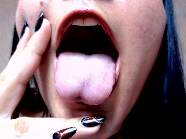 NaniSky's open mouth with tongue out, pink tongue hanging, black nail polish, hand with same nail polish touching bottom lip, dark hair, light skin, slightly glossy lips.