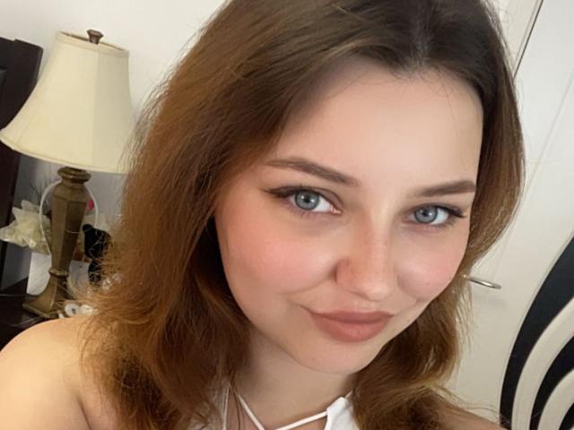 LilSoft tiene ojos azules, cabello ondulado de color castaño claro y una sonrisa sutil. Lleva una blusa blanca, y sus senos son ligeramente visibles. Se ve linda y sexy..