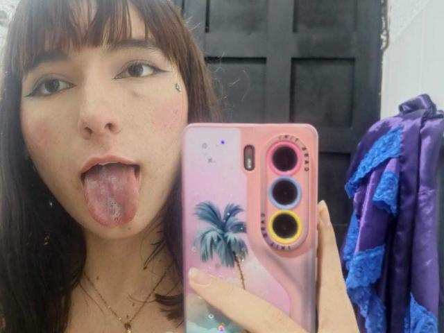 HairyValeri met bruin haar en sproeten, met haar tong uit, houdt een roze telefoon vast met een palmboom ontwerp, met drie camera lenzen, in een spiegel-selfie. Haar achterkant is niet zichtbaar..