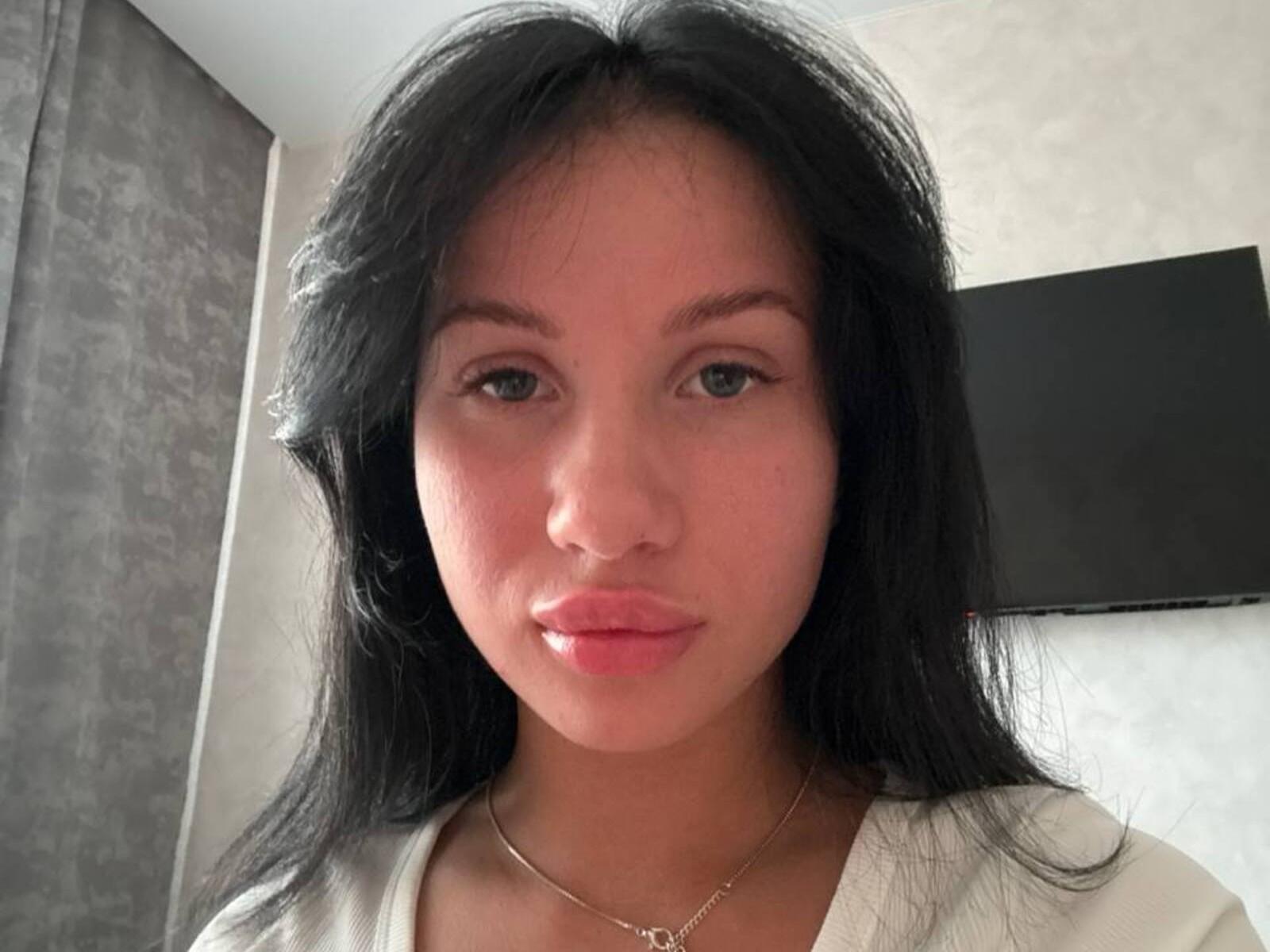 Sunshine Amendola hat langes, schwarzes Haar, hellbraune Haut, blaue Augen und glänzende, rosafarbene Lippen. Sie trägt ein weißes Oberteil und eine goldene Halskette und blickt direkt in die Kamera..