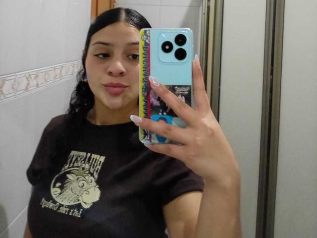 LonxEva with black hair, wearing a brown shirt, holding a blue phone with colorful case, taking a mirror selfie, showing pouty lips, and visible boobs in the reflection.