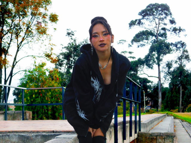 HenutmireRossi leans forward, wearing a black hoodie with white patterns, dark lipstick, and a necklace, standing on stone steps with a metal railing in a wooded area.