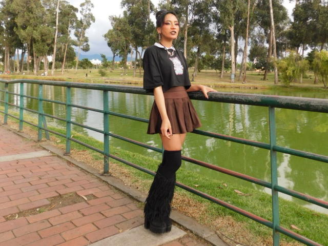 HenutmireRossi stands by a green railing, wearing a black top, brown skirt, black knee socks, and black shoes, with her hand on the railing, and trees in the background.