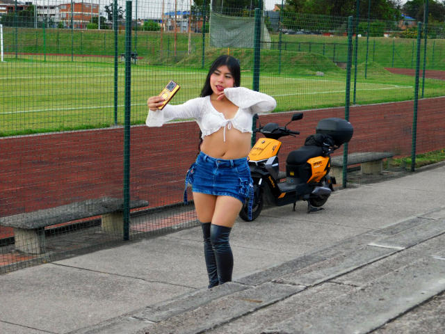 CataMoreno with black hair, white tied-up crop top, blue denim skirt, black knee-high boots, holding phone, standing next to yellow scooter, smiling, outdoor, daytime.