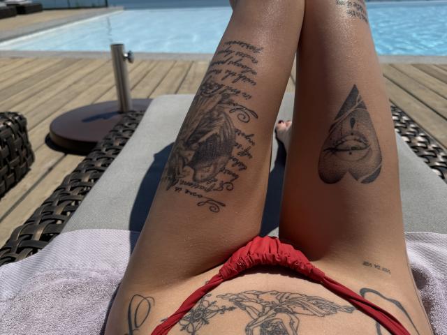 SensualMila's red bikini bottom, tattooed legs on a lounge chair, showing a heart tattoo on her right thigh and a face tattoo on her left leg, by a pool.