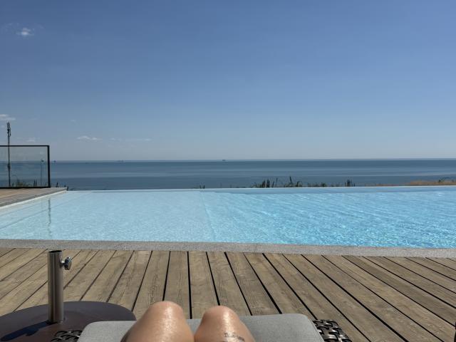 SensualMila's wet thighs and knees are in the bottom foreground, with a blue infinity pool and a calm sea under a clear sky in the background.