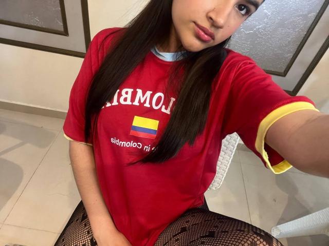 IrisBlair wearing a red shirt, fishnet pants, and pigtails, with a small Colombian flag on her shirt, sitting with her hand near her pussy. She has a cute face.