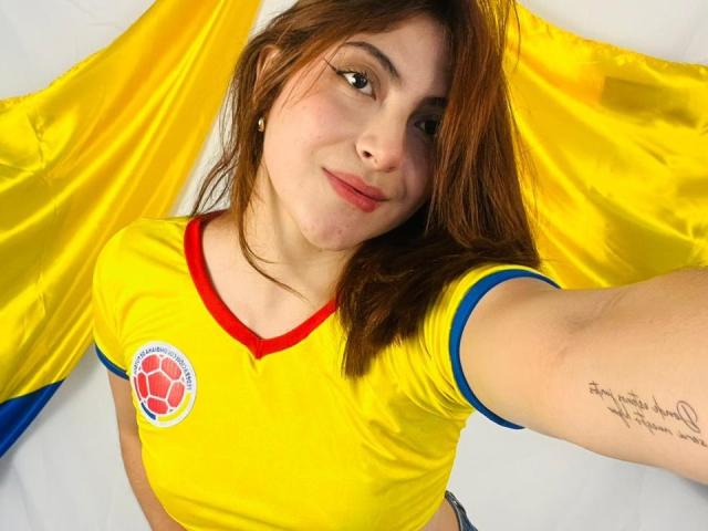 SpencerLe, avec des cheveux bruns, un maillot de foot jaune avec des détails rouges et bleus, un logo de ballon de foot, souriant, avec un tatouage sur le bras, des drapeaux jaunes en arrière-plan, tenant un téléphone pour un selfie..