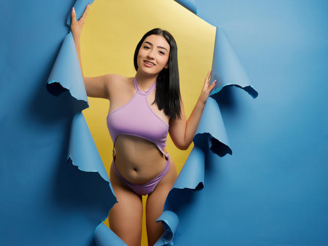 ZoeyMorelli a de grandes seins, elle porte un haut et une culotte assortis de couleur lavande, et elle sourit joyeusement, les bras tenant du papier bleu et jaune sur un fond bleu uni..