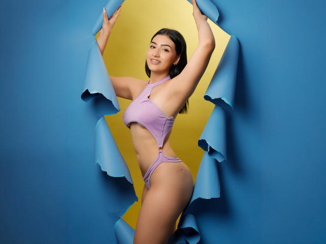 Zoey Morelli mit einem blasslila Bikini, Armen erhoben, stehend in einem blauen und gelben Loch, lächelnd, wobei ihr Hintern zu sehen ist, kleine Brüste, süß, dunkles Haar, helle Haut, schlank, sexy..