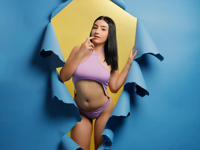 Zoey Morelli, con capelli neri, reggiseno sportivo viola e mutandine abbinate, con un dito in bocca, su uno sfondo di carta blu e giallo, che mostra un leggero girovita e un sedere piccolo..