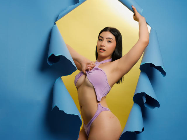 Zoey Morelli in een lila badpak met uitsparingen, die de bovenkant vasthoudt, waarbij haar kleine borsten en platte buik zichtbaar zijn, met armen omhoog, donker haar, en een achtergrond in blauw en geel..