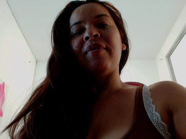 LizethBells met donkerbruin haar, die lichtjes glimlacht, draagt een bruin tanktopje met kantrand, waardoor haar kleine borsten zichtbaar zijn, en heeft donkere ogen, op een selfie genomen vanuit een lage hoek..