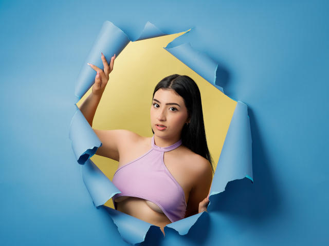 Zoey Morelli, mit langen schwarzen Haaren, trägt ein pinkes Halstopp und lugt durch ein blaues Papierloch. Dabei sind ihre Brüste und ein gelber Hintergrund zu sehen. Sie hat ein süßes, sexy Aussehen..