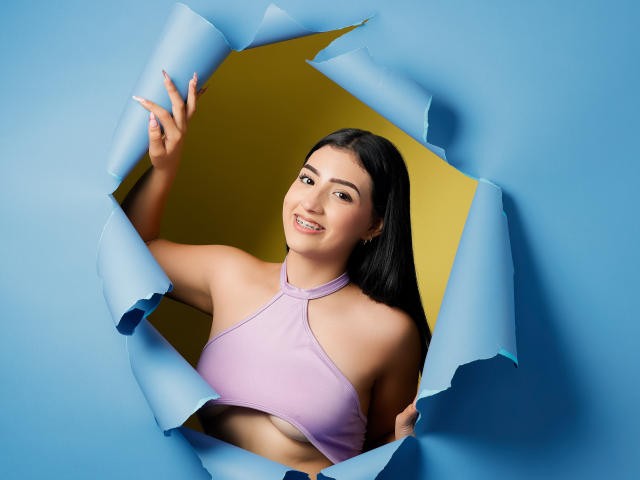 Zoey Morelli, avec de longs cheveux noirs, souriante, portant un haut rose à encolure dégagée, ses seins légèrement visibles, regardant à travers un trou en papier bleu, fond jaune, main droite levée..
