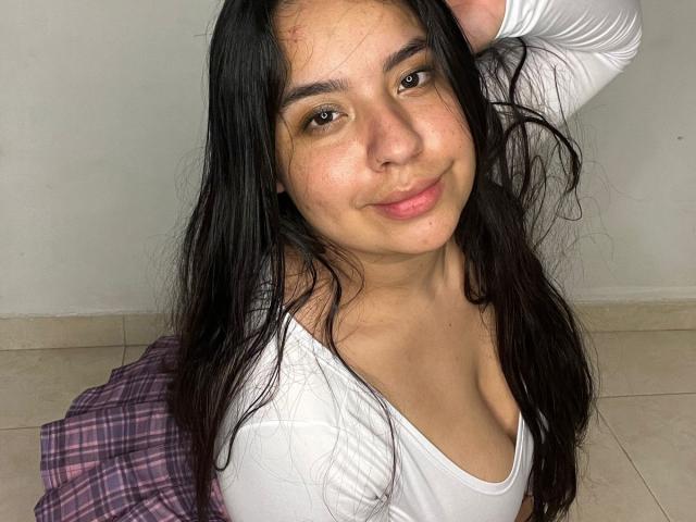 SammyTitss with long black hair, brown eyes, and a slight smile, wearing a white top, showing her small tits, sitting on a tiled floor with a purple plaid shirt.