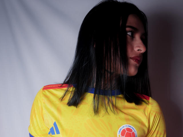 NicolleSweetty, con i capelli neri a metà schiena, indossa una maglia da calcio gialla con strisce rosse e blu, guarda di lato, mostrando il suo profilo e il rossetto rosso..