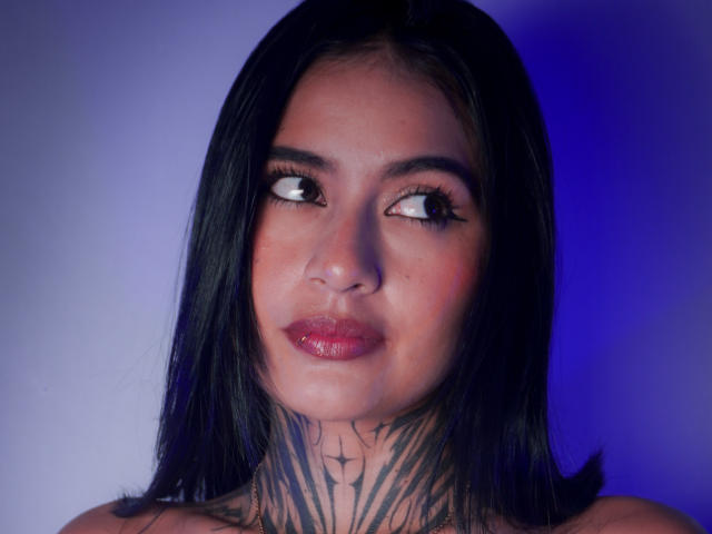 NicolleSweetty tem cabelo preto, lábios vermelhos e tatuagens no pescoço. Ela olha para o lado com um leve sorriso. Seus olhos são escuros e seu rosto está brilhante..