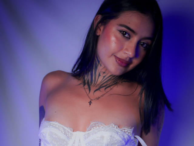 NicolleSweetty, con lunghi capelli neri, sorridente, indossa un top bianco in pizzo senza spalline, mostrando tatuaggi sul collo e sulla parte superiore del petto, illuminazione blu e viola, leggero rossore sulle guance, una piccola collana con una croce..