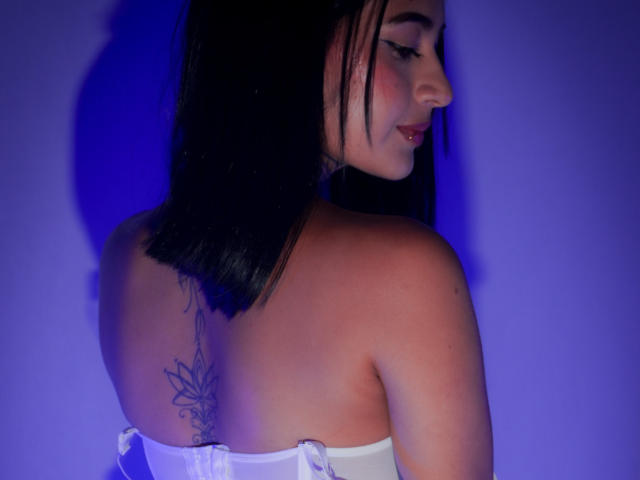 NicolleSweetty mit dunklem, schulterlangem Haar, einem weißen Top ohne Träger, das ein Tattoo auf ihrem Rücken zeigt, die über ihre Schulter schaut, blaues Licht, freie Schultern, ein leichtes Lächeln, süß..