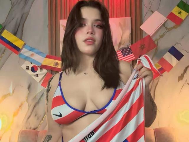 AryButty with medium tits, wearing a red, white, and blue bikini top, holding a flag, has dark hair, red eyeshadow, and tattoos, surrounded by flags.