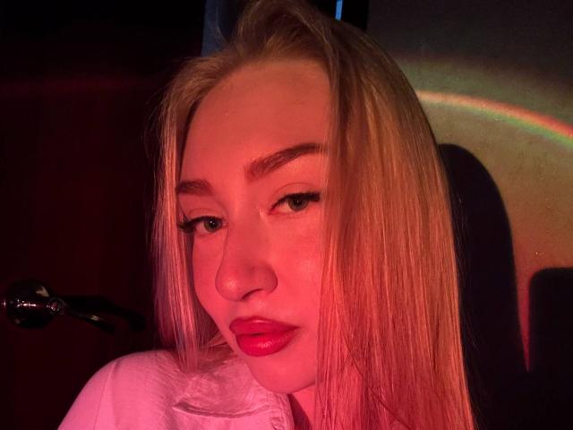 JoetteShigeta with long blonde hair, red lipstick, and subtle eye makeup, looking slightly down with a relaxed expression, illuminated by pink and red lighting.