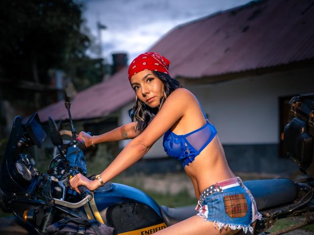 EvelinLuna mit blauem BH, Jeansshorts, rotem Bandana, auf einem blauen Motorrad sitzend, zeigt ihr trainiertes Gesäß und ihre kleinen Brüste, sieht sexy aus, Dämmerlicht, Tattoo am rechten Arm..