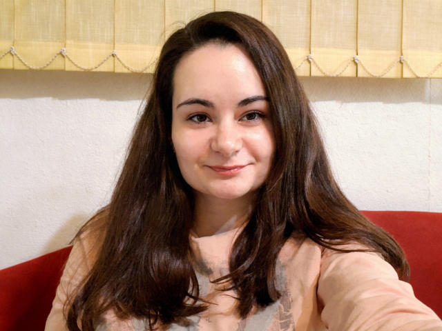 ElizanXStarzy tiene el pelo castaño de longitud media, piel clara y una sonrisa sutil. Lleva una blusa larga color melocotón que deja ver sus pequeños senos. En el fondo, hay un sofá rojo..
