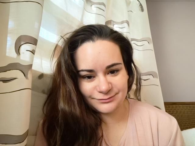 ElizanXStarzy, con lunghi capelli castani, pelle chiara e un sorriso delicato, indossa una camicia rosa ed è in piedi di fronte a tende a motivi. Il suo seno è piccolo e non visibile..