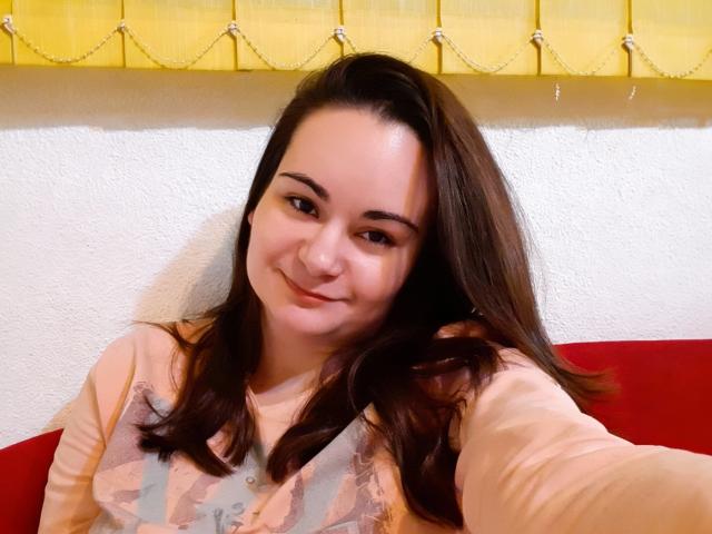 ElizanXStarzy mit mittelgroßer Brust, lächelnd, braunes Haar, trägt ein rosa und graues gemustertes Hemd, macht ein Selfie, gelb vorhänge im Hintergrund, weiße, strukturierte Wand, rotes Kissen..