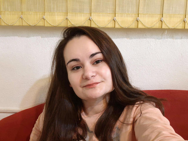 ElizanXStarzy, con lunghi capelli castani, pelle chiara e un sorriso delicato, indossa una maglietta rosa, seduta su un divano rosso, con delle persiane gialle sullo sfondo..
