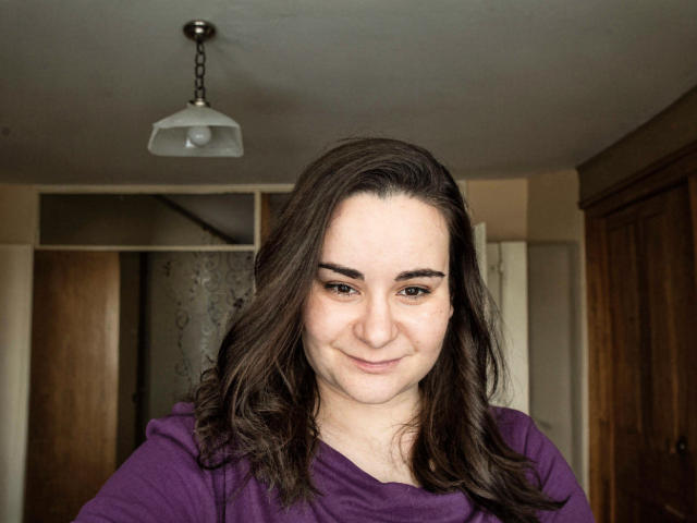 ElizanXStarzy, mit welligem, braunem Haar, einem lila Hemd und einem leichten Lächeln, hat dunkle Augenbrauen und helle Haut und steht in einem Raum, in dem eine Hängelampe hängt..