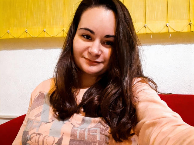 ElizanXStarzy, con cabello largo y castaño oscuro, sonriendo, vistiendo una camisa con estampado rosa y marrón, selfie, cortina amarilla, pared blanca y sofá rojo en el fondo..