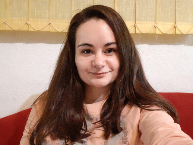 ElizanXStarzy ha capelli castano di media lunghezza, una carnagione chiara e sta sorridendo leggermente. Indossa una camicia color pesca ed è seduta su un cuscino rosso..