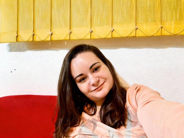 ElizanXStarzy, con lunghi capelli castani, sorridente, indossa una camicia rosa, è seduta contro un cuscino rosso, con una tenda gialla sullo sfondo, una parete bianca con texture e piccole macchie scure sul viso..