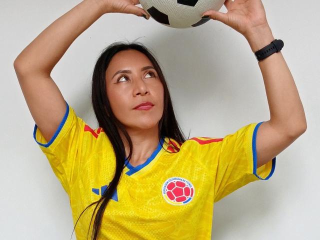 VivienneWest, con cabello negro largo, sosteniendo una pelota de fútbol por encima de la cabeza, vistiendo una camiseta de fútbol amarilla con detalles azules y un emblema rojo, mirando hacia arriba, y usando un reloj de pulsera negro..