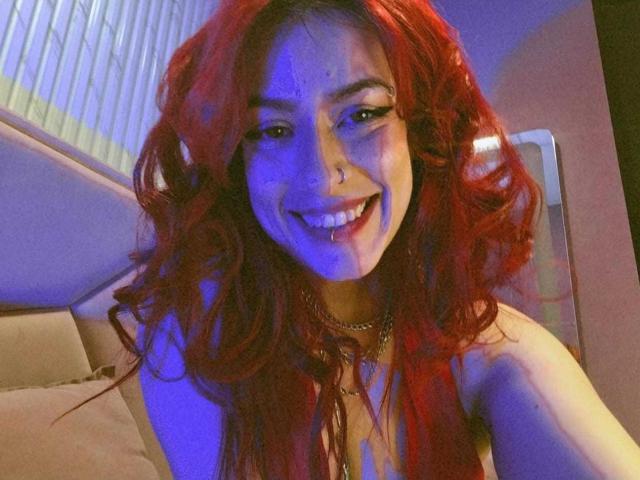 LolyYunno, con cabello rizado rojo, sonriendo, con luces azules y rosas en su rostro, piercing en la nariz, piercing en el labio, collar y una blusa sin mangas, que muestra ligeramente sus senos en la foto..