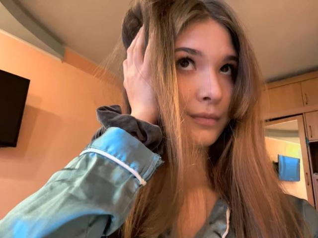 JanyceCastlen, con cabello castaño largo, sosteniendo una goma para el pelo en su mano derecha, vistiendo una chaqueta verde, mirando a la cámara con una ligera sonrisa, en una habitación bien iluminada..