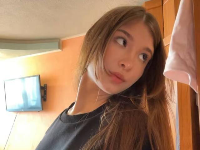 JanyceCastlen, con cabello castaño largo, nariz pequeña y maquillaje discreto, vestida con una camisa negra, mirando por encima del hombro con un ligero ceño fruncido, en una habitación de madera..