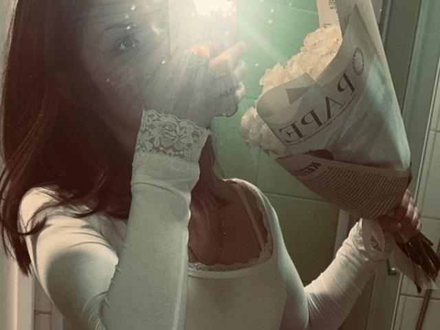 JanyceCastlen, con i capelli castani, le maniche di pizzo bianco e la maglietta bianca, tiene in mano un sacchetto di patatine, coprendosi la bocca con la mano, con riflessi solari sul viso, con il seno visibile..