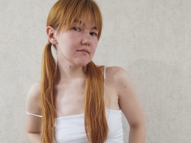 SheritaChieng with red pigtails, wearing a white spaghetti-strap top, has small tits, a fair skin, and a slight smile, against a plain, textured wall.