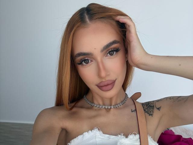 MarissaFex, avec de longs cheveux orange, portant un haut blanc en dentelle, un collier ras du cou et un tatouage sur l'épaule droite, une main dans les cheveux, regardant la caméra avec un maquillage complet et des lèvres pulpeuses..