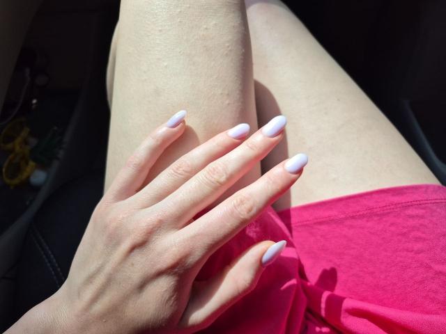 Les jambes pâles de Rozalin, avec une chemise rose, sa main aux ongles lavande reposant sur sa cuisse. La lumière du soleil met en valeur sa peau claire. Ses doigts sont fins, les ongles longs et pointus..