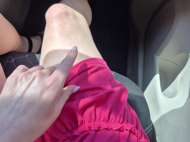 Rozalins knie bevindt zich in een roze jurk, een hand met lichtblauwe nagellak wijst ernaar, en toont haar dijbeen op de grijze stoel van een auto..