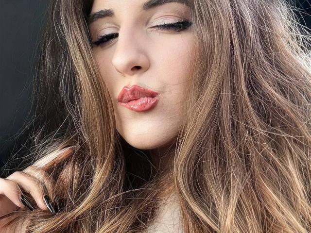 Maisie Vandenberghe, com lábios rosados e curvados, olhos fechados e longos cabelos castanhos ondulados, tocando o cabelo com unhas pretas e bem cuidadas, com uma aparência sexy e sedutora..