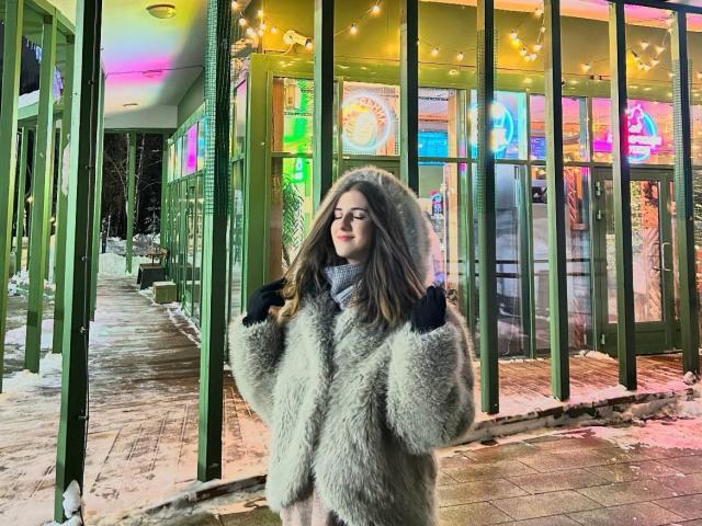 Maisie Vandenberghe steht in einem schneebedeckten, neonbeleuchteten Bereich und trägt einen flauschigen, grauen Kapuzenmantel, schwarze Handschuhe und eine Brille. Sie hat einen ruhigen Gesichtsausdruck und hält die Hände in den Taschen ihres Mantels..
