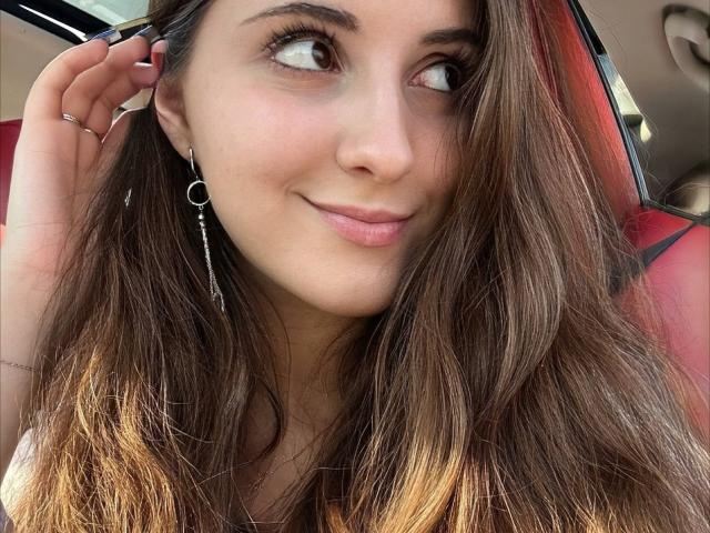 Maisie Vandenberghe, con lunghi capelli castani, occhi castani e un leggero sorriso, indossa un orecchino d'argento, lo tocca con le dita, è seduta in una macchina rossa e mostra un seno carino e piccolo..